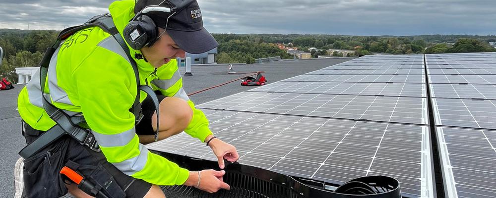 En montör från Nordic Solar monterar nya solceller och fågelskydd på ett stort fastighetstak. Arbetet sker med säkerhetssele, och panelerna läggs i jämna rader för maximal effekt. Pressbild: Nordic Solar