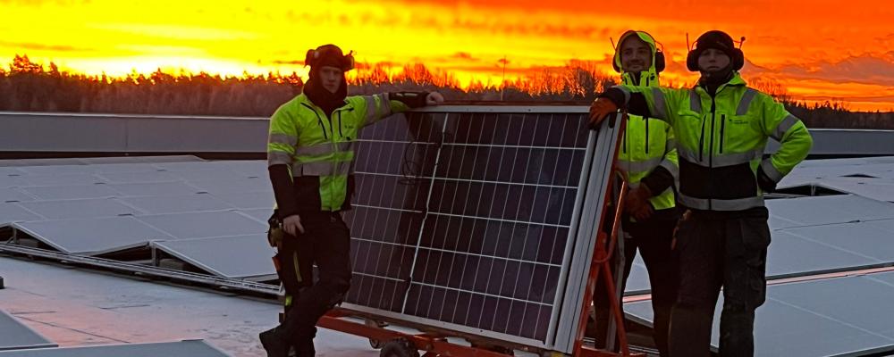 Bilden visar tre montörer som arbetar med installationen av solpaneler på taket vid Solar Sveriges logistikcentrum. De står bredvid en vagn med solpaneler medan himlen bakom dem lyser i intensiva orangea och röda toner från solnedgången. Pressbild: Nordic Solar