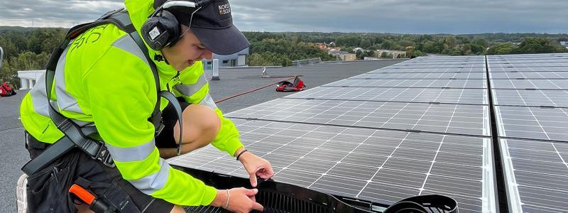 En montör från Nordic Solar monterar nya solceller och fågelskydd på ett stort fastighetstak. Arbetet sker med säkerhetssele, och panelerna läggs i jämna rader för maximal effekt. Pressbild: Nordic Solar