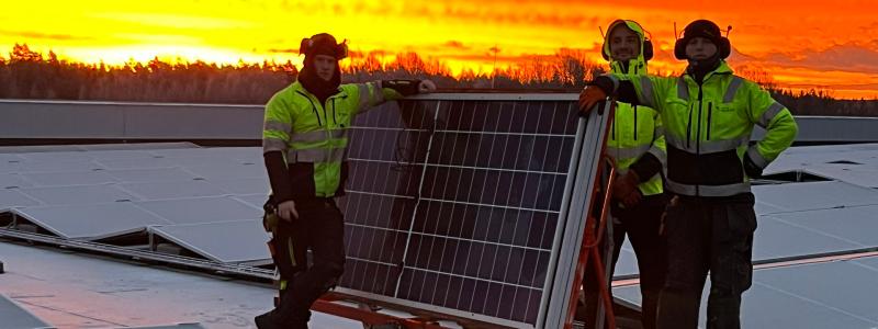 Bilden visar tre montörer som arbetar med installationen av solpaneler på taket vid Solar Sveriges logistikcentrum. De står bredvid en vagn med solpaneler medan himlen bakom dem lyser i intensiva orangea och röda toner från solnedgången. Pressbild: Nordic Solar