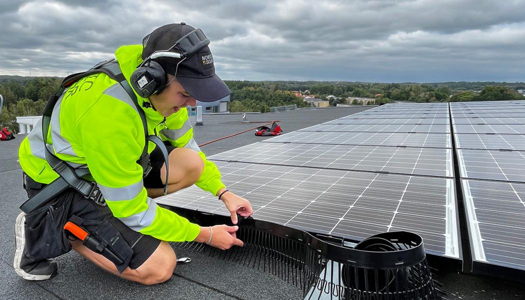 En montör från Nordic Solar monterar nya solceller och fågelskydd på ett stort fastighetstak. Arbetet sker med säkerhetssele, och panelerna läggs i jämna rader för maximal effekt. Pressbild: Nordic Solar