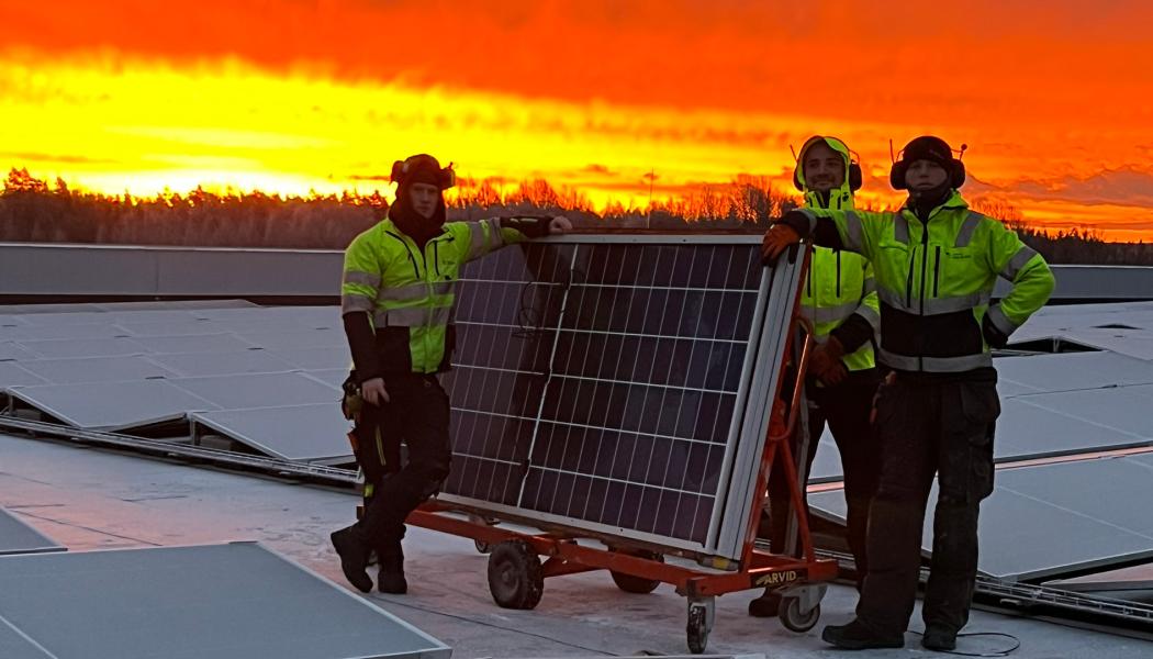 Bilden visar tre montörer som arbetar med installationen av solpaneler på taket vid Solar Sveriges logistikcentrum. De står bredvid en vagn med solpaneler medan himlen bakom dem lyser i intensiva orangea och röda toner från solnedgången. Pressbild: Nordic Solar