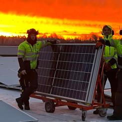 Bilden visar tre montörer som arbetar med installationen av solpaneler på taket vid Solar Sveriges logistikcentrum. De står bredvid en vagn med solpaneler medan himlen bakom dem lyser i intensiva orangea och röda toner från solnedgången. Pressbild: Nordic Solar